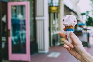 A person's hand holding a pink ice cream cone in front of a shop with a pink door.